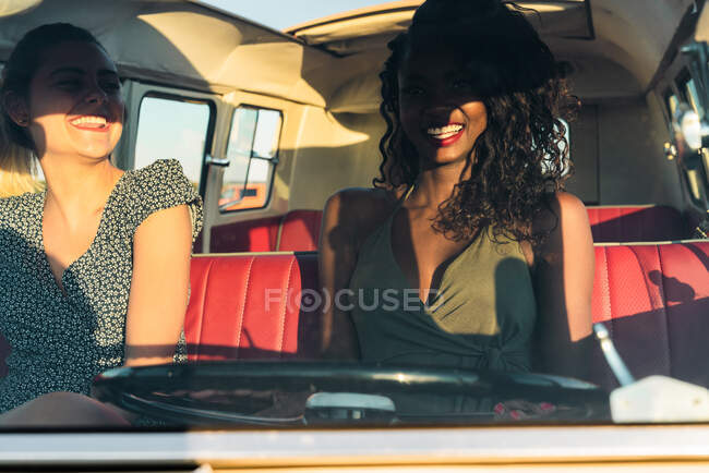 Deux jeunes femmes assises à l'intérieur d'un van vintage et souriant tout en voyageant dans la nature ensemble — Photo de stock