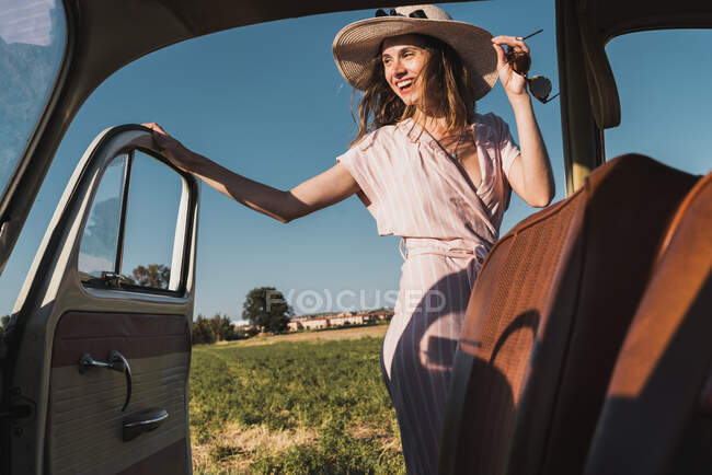 Vista desde el interior del coche retro de la mujer de moda feliz en sombrero y gafas de sol de pie con entusiasmo en la naturaleza y riendo - foto de stock