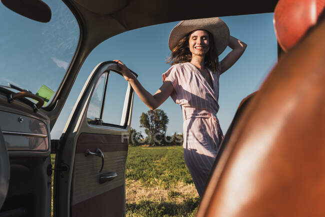 Vue de l'intérieur de la voiture rétro de la femme heureuse à la mode dans le chapeau et les lunettes de soleil debout avec enthousiasme dans la nature et en riant — Photo de stock