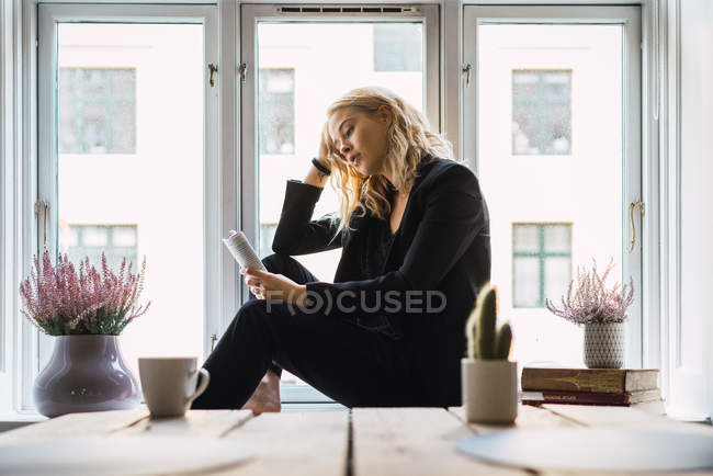 Träumende Frau liest Buch, während sie zu Hause auf der Fensterbank sitzt — Stockfoto