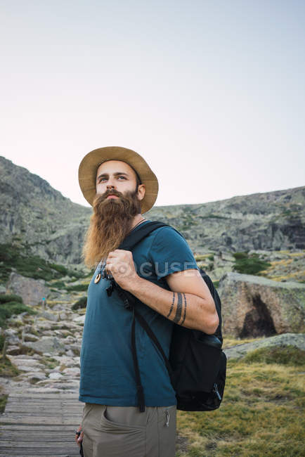 Молодой человек в шляпе с рюкзаком, стоящим у подножия горы — стоковое фото