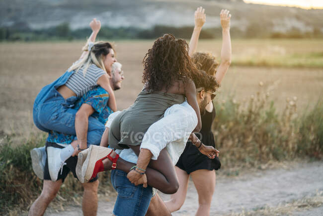 Two men giving piggyback ride to women and running along countryside road while spending time in nature together — Stock Photo