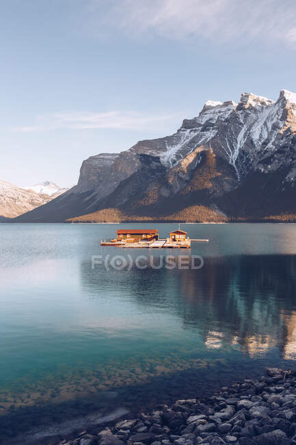 Будівля з коричневим дахом під сонцем посеред високогірного озера з прозорою блакитною водою на фоні мальовничих гір та лісу — стокове фото