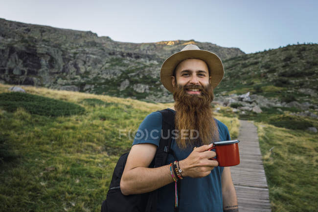 Junger Mann steht mit Becher auf Pfad in den Bergen und blickt in Kamera — Stockfoto