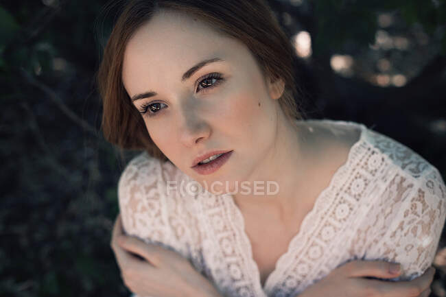 From above shot of pretty young lady in lace apparel embracing herself and looking away while standing on blurred background of nature — Stock Photo