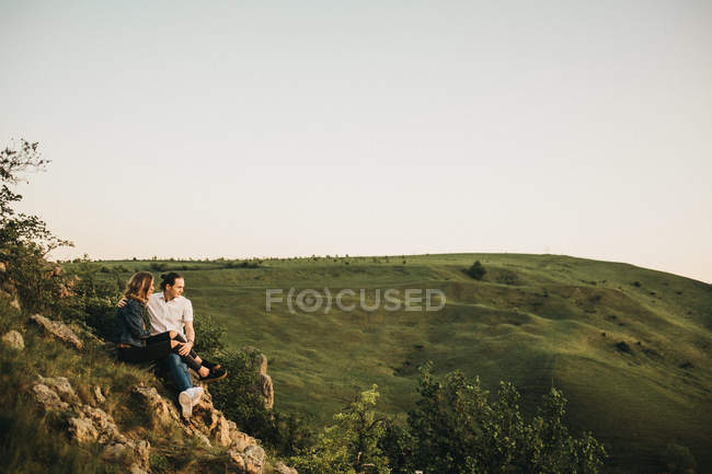 Симпатична пара обіймається, сидячи на скелястому схилі на тлі красивої долини і гір — стокове фото