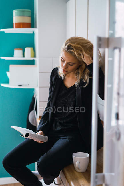 Mujer relajada casual tomando café mientras está sentada en el mostrador de la cocina y leyendo libro - foto de stock