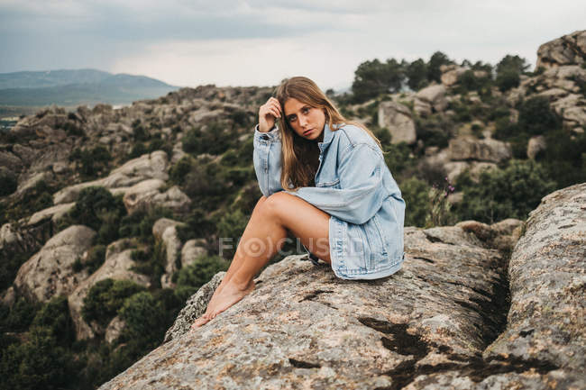 Молода жінка в джинсовій куртці сидить наодинці на величезній скелястій скелі в природі дивлячись на камеру — стокове фото