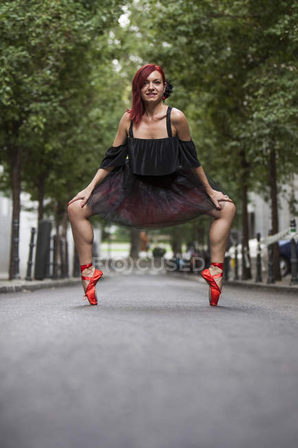 Балерина червоної голови з чорним навчальним посібником та червоним балетом танцює на вулиці з деревами на задньому плані . — стокове фото