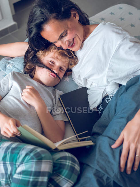 Carino ragazzo avendo divertente mentre sdraiato su letto e lettura interessante libro con aiuto di sorridente madre — Foto stock