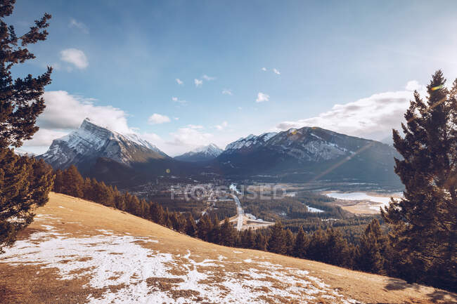 Hill with yellow frozen soil and thick forest on background with snowy mountains and blue sky with few clouds and valley with small city — Stock Photo