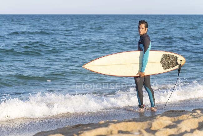 Человек с доской для серфинга стоит на мокром песчаном пляже у океана
. — стоковое фото