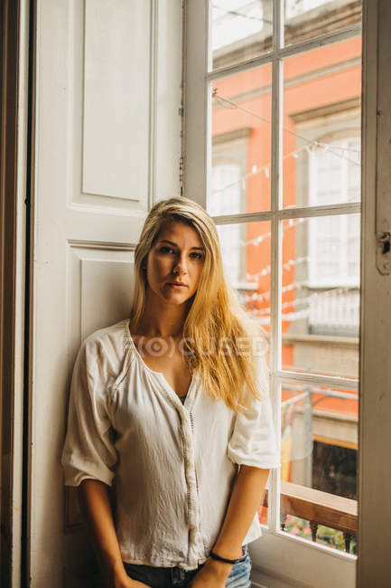 Портрет молодой женщины в повседневной одежде, смотрящей в камеру, стоя у окна в уютной комнате — стоковое фото