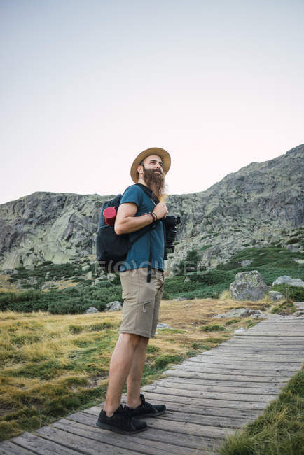 Young bearded man in hat with backpack standing on wooden way leading to mountains — Stock Photo