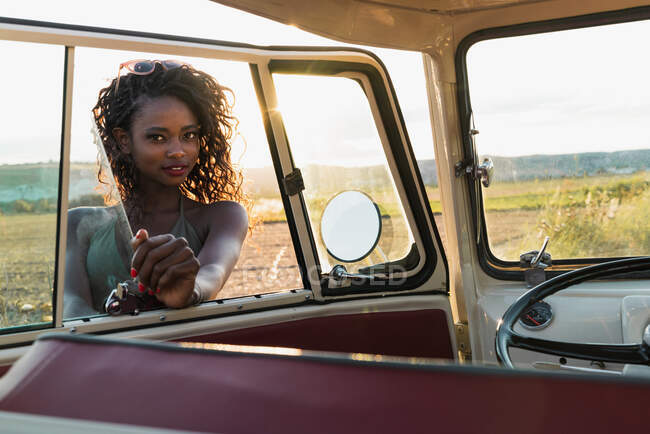 Preciosa mujer afroamericana sonriendo y mirando a la cámara a través de la ventana del coche vintage mientras pasa tiempo en la naturaleza en el día soleado - foto de stock