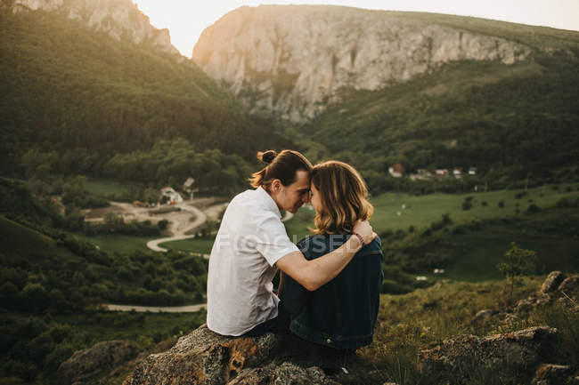 Couple mignon embrassant et endurcissant les fronts tout en étant assis sur une pente rocheuse sur fond de belle vallée et de montagnes — Photo de stock