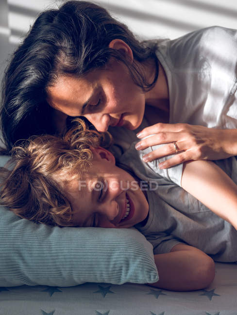Hermosa mujer acostada en la cama detrás de dulce chico y tocando su mejilla cuidadosamente - foto de stock