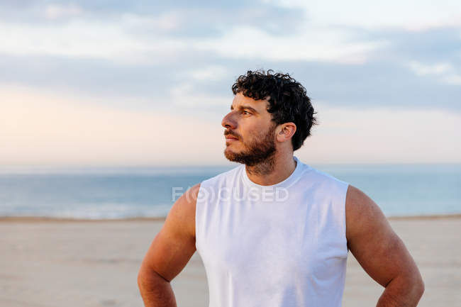 Uomo positivo in abbigliamento sportivo con le mani in vita mentre in piedi sulla spiaggia di sabbia durante il tramonto — Foto stock