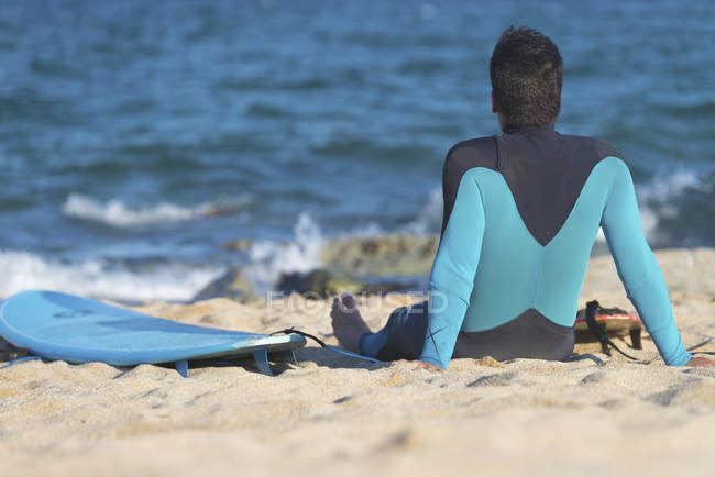 Vista trasera del hombre en traje de neopreno sentado con tabla de surf en la playa mirando al océano . - foto de stock