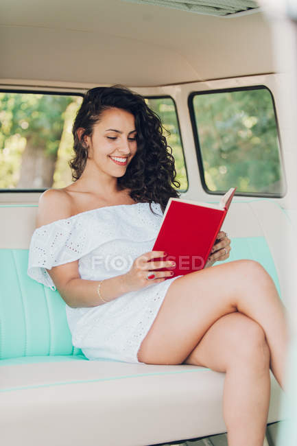 Sonriente joven sentada dentro de la caravana y leyendo el libro - foto de stock