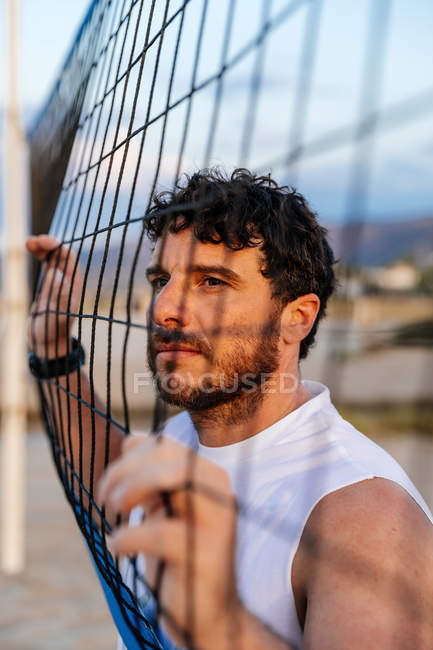 Uomo barbuto in abbigliamento sportivo toccare rete da pallavolo e distogliere lo sguardo durante l'allenamento sulla spiaggia — Foto stock