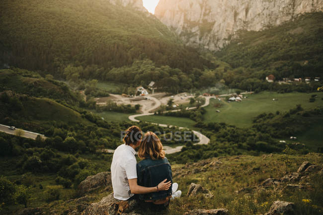 Nettes Paar umarmt und strafft die Stirn, während es auf einem felsigen Hang vor dem Hintergrund des schönen Tales und der Berge sitzt — Stockfoto