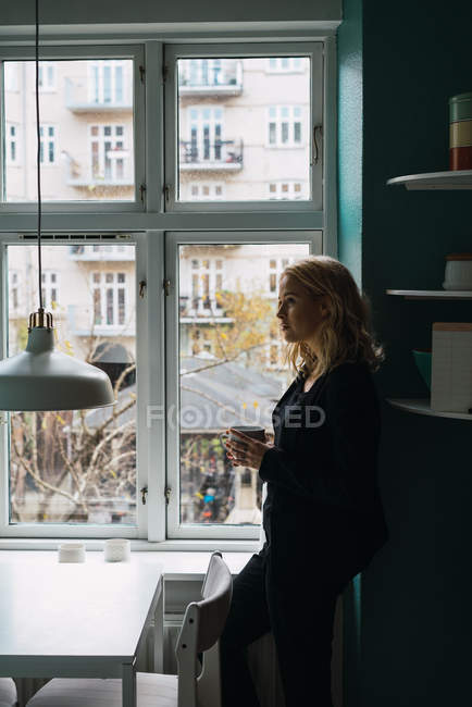 Donna romantica con tazza di bevanda in piedi vicino alla finestra a casa — Foto stock