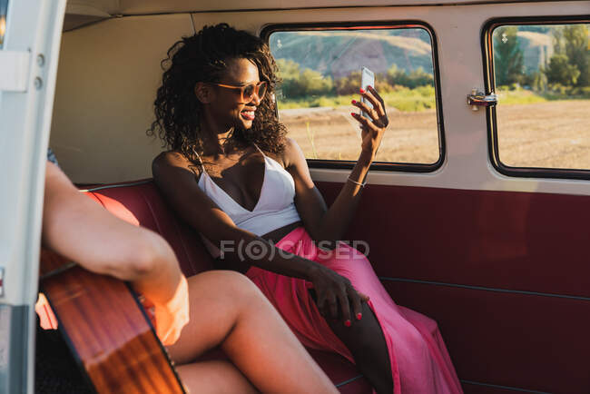 Diversas mujeres tocando la guitarra acústica y riendo mientras se sientan juntos dentro de van retro durante el viaje - foto de stock