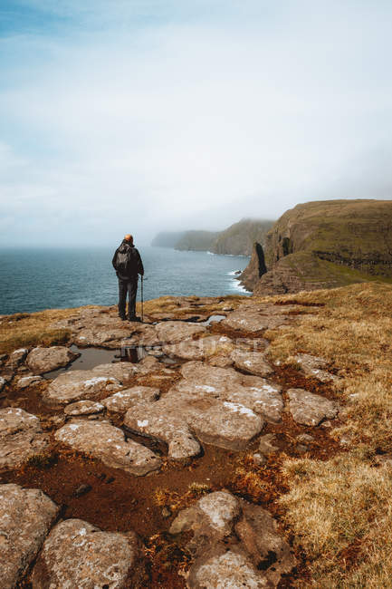 Caminante irreconocible de pie en la costa con palo y mirando al océano en las Islas Feroe - foto de stock