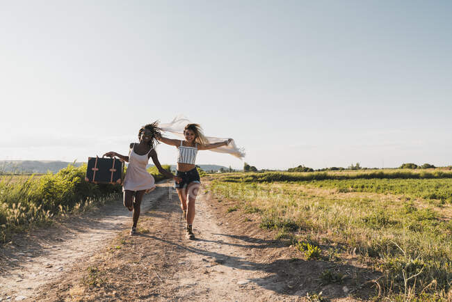 Веселі стильні багатоетнічні жінки з валізою і шарфом з ентузіазмом бігають по дорозі в літній зеленій сільській місцевості — стокове фото
