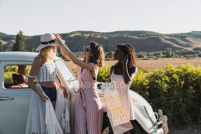 Модні багатоетнічні жінки, що стоять біля старовинного автомобіля і читають карту під час подорожей разом влітку — стокове фото