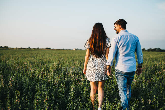 Cheerful young man and woman holding hands and walking on field in sunlight — Stock Photo