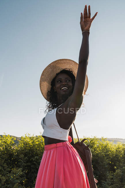 Trendy African-American woman in hat carrying suitcase and walking on rural road in summertime — Stock Photo