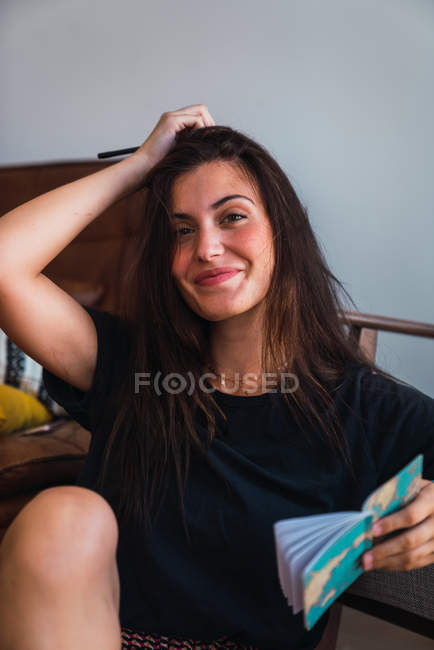 Jovem mulher morena sorridente em camiseta preta sentada no quarto com notebook e olhando para a câmera — Fotografia de Stock