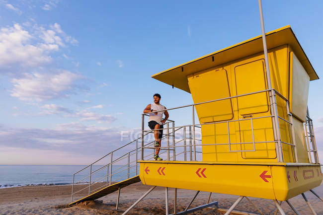 Бородатый мужчина в спортивной одежде, опирающийся на перила хижины спасателя на пляже во время тренировки на открытом воздухе на закате — стоковое фото