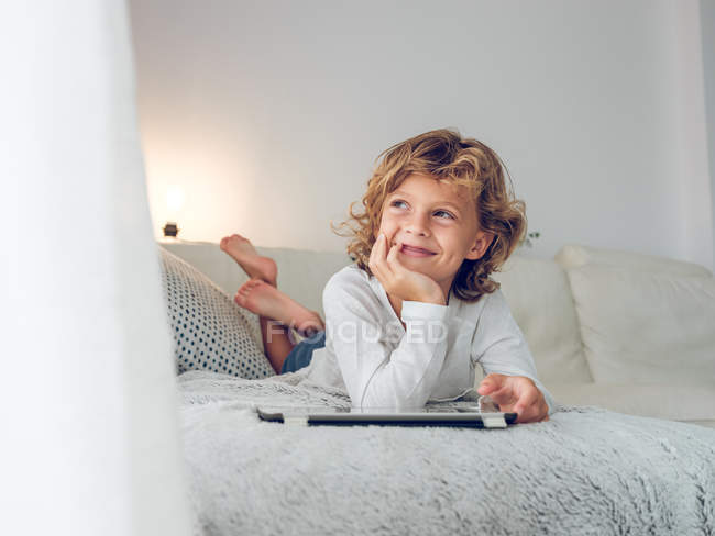 Fröhlicher Junge liegt mit digitalem Tablet auf Couch und schaut weg — Stockfoto