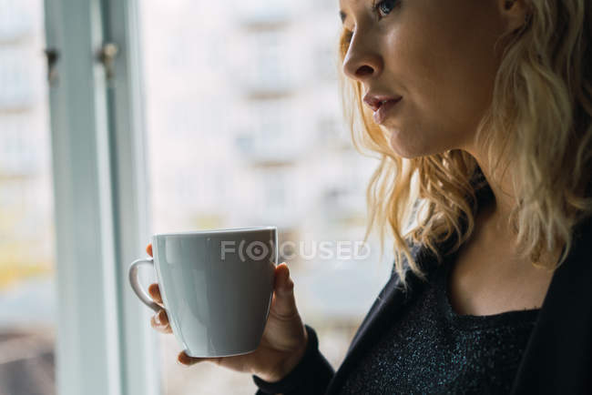 Primer plano de la joven reflexiva sosteniendo taza de bebida en casa - foto de stock