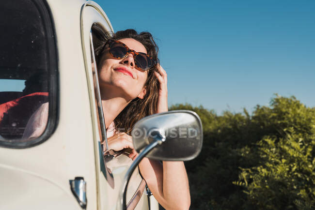 Jovem mulher feliz em óculos de sol elegantes olhando para fora da janela do carro desfrutando de luz solar de verão contra árvores verdes e céu azul — Fotografia de Stock