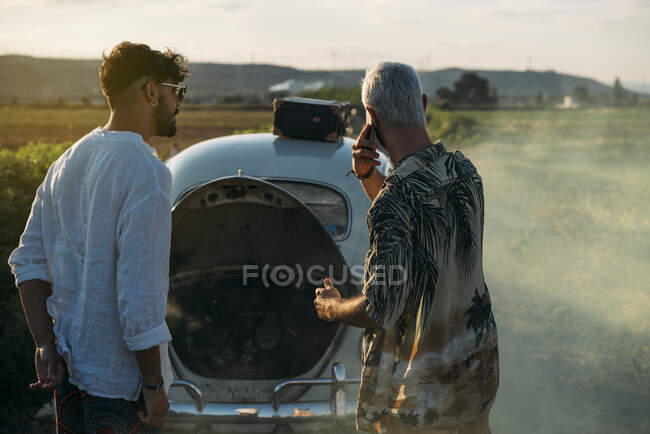 Handsome bearded man having smartphone conversation while standing on countryside road near male friend and broken car emitting thick fume — Stock Photo
