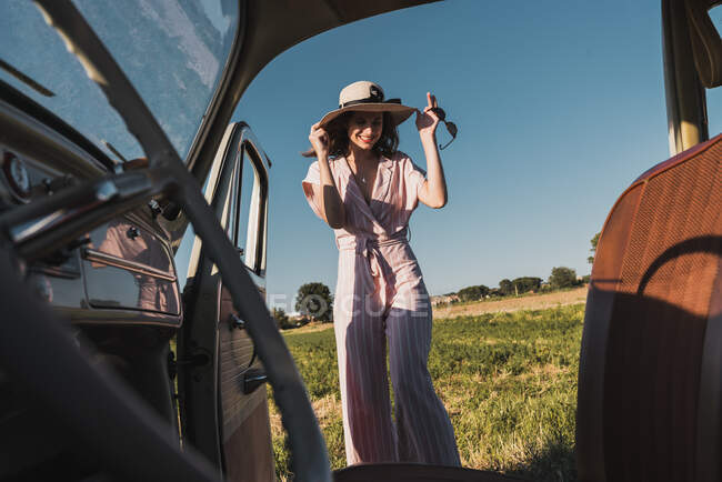 Vista desde el interior del coche retro de la mujer de moda feliz en sombrero y gafas de sol de pie con entusiasmo en la naturaleza y riendo - foto de stock