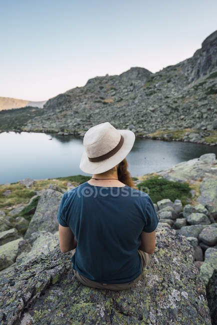 Junger Mann mit Hut sitzt auf Felsen in der Nähe des Sees und blickt auf die Aussicht — Stockfoto