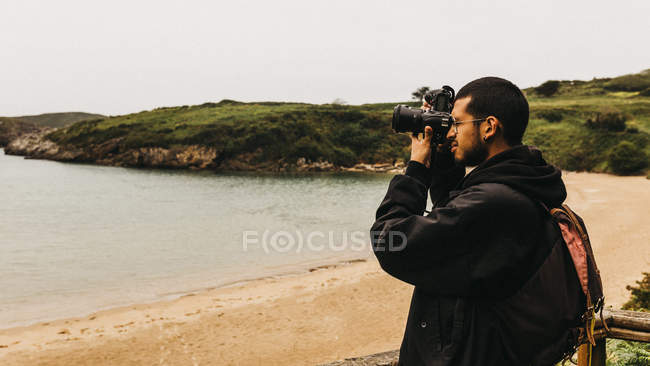 Vista lateral de chico guapo con mochila de pie en la costa de arena y tomar fotos de hermoso océano durante el viaje en la naturaleza - foto de stock
