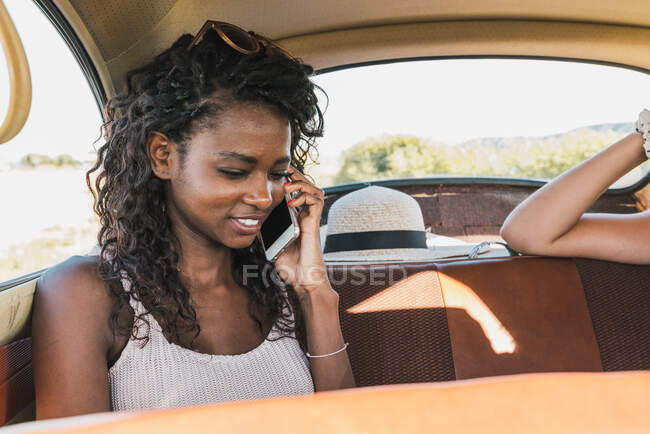Молода афро-американська жінка, яка телефонує, сидячи на задньому сидінні старої машини з мандрівними друзями. — стокове фото