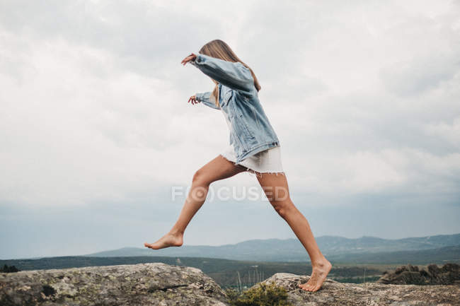 Боковой вид женщины в платье и джинсовой куртке, прыгающей через крэк, прыгающей со скалы на скале против облачного неба — стоковое фото
