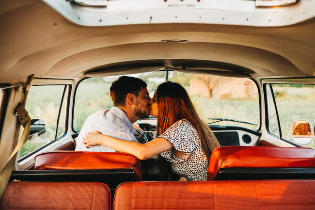 Vista trasera de hombre y mujer jóvenes sentados en furgoneta vintage y besándose en la naturaleza - foto de stock