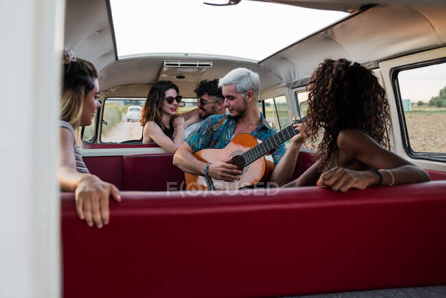 Group of young people laughing and listening to handsome guy playing acoustic guitar inside vintage van in nature — Stock Photo