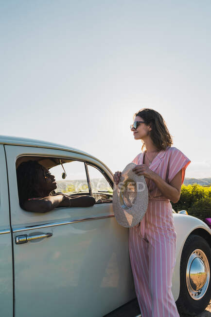 Стильная брюнетка в солнцезащитных очках, опирающаяся на автомобиль снаружи и беседующая с симпатичной черной женщиной внутри при ярком солнечном свете летом — стоковое фото
