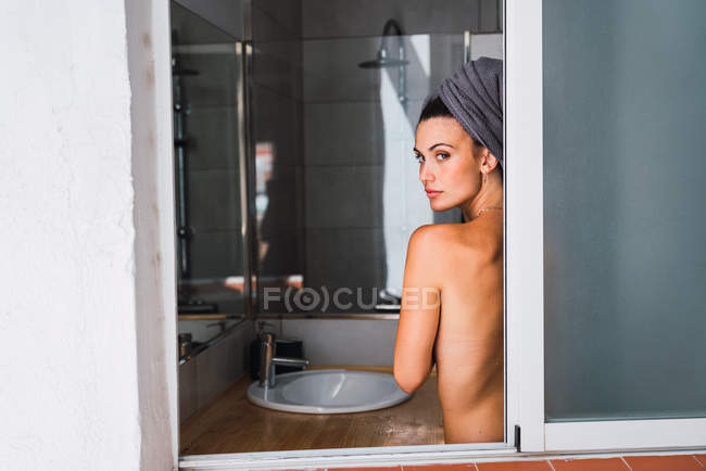 Молодая топлесс женщина, стоящая в ванной комнате с полотенцем на голове — стоковое фото