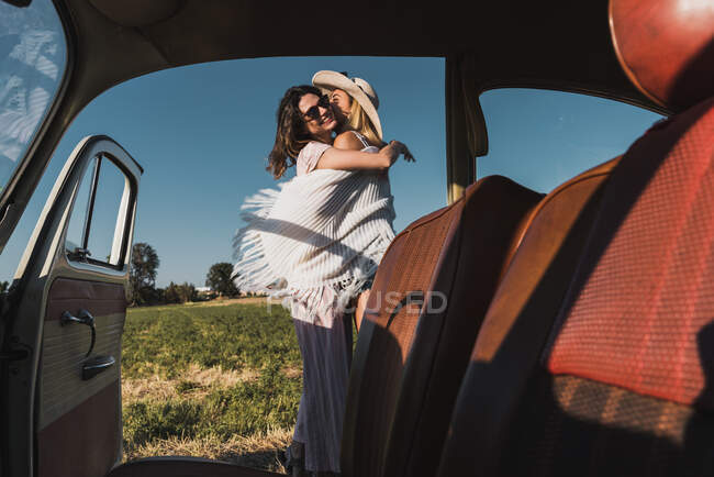 Vista de dentro do carro retro de mulheres abraçando e beijando alegremente fora contra a paisagem com árvores verdes e céu — Fotografia de Stock