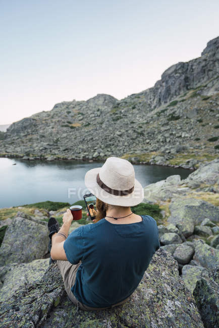 Молодой человек сидит на скалах возле озера с чашкой и с помощью смартфона — стоковое фото